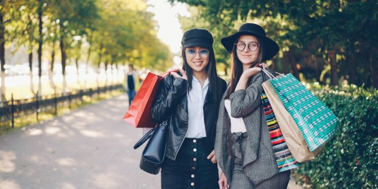 Portrait of happy girls shopaholics standing outdoors with bright bags, looking at camera and smiling touching hair. Shopping, people and modern youth lifestyle concept.