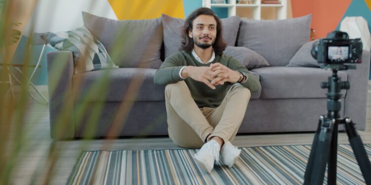 Happy Arab man is recording video for internet blog speaking and gesturing sitting on floor at home. Vlogging, youth and modern technology concept.